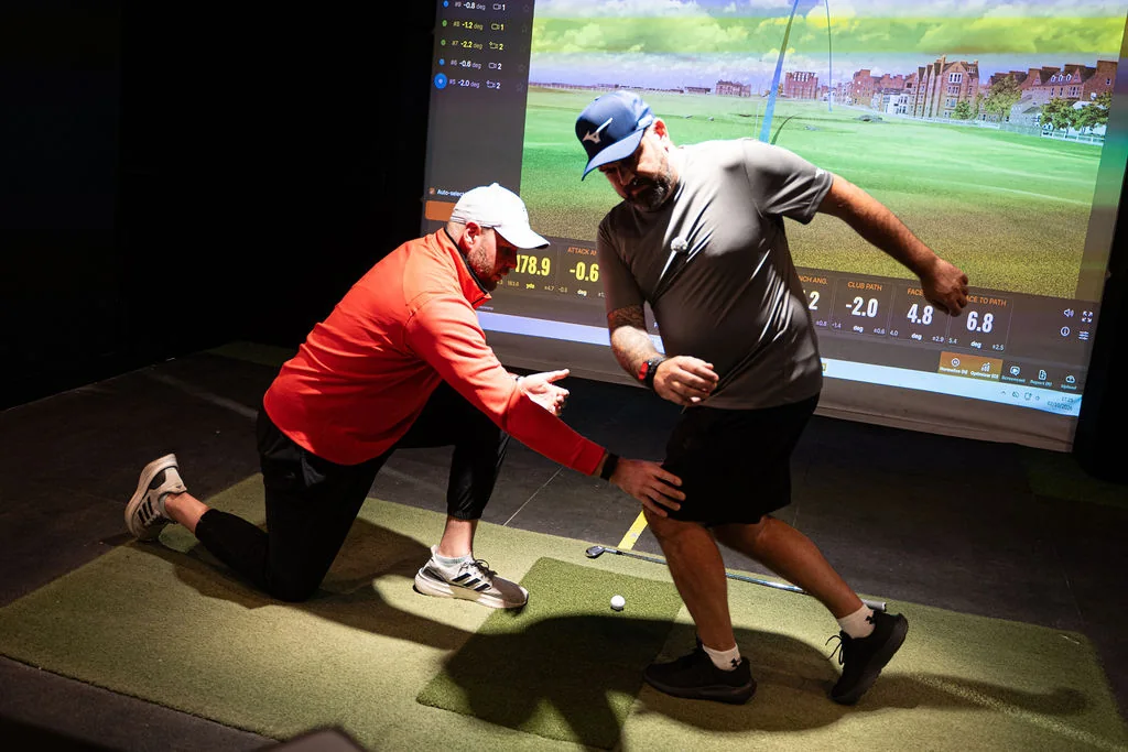 Coach kneeling beside a golfer during a balance and movement drill in the TrackMan bay at Evolution Golf Academy near Derby