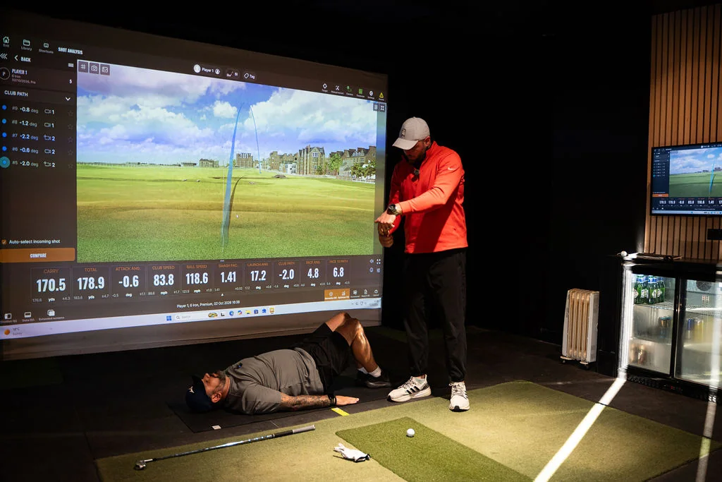 Golfer lying on the mat during a floor mobility assessment with the coach beside the TrackMan screen at Evolution Golf Academy near Derby