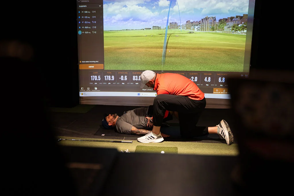 Coach assessing hip mobility with the golfer lying on the mat in front of the TrackMan screen at Evolution Golf Academy near Derby
