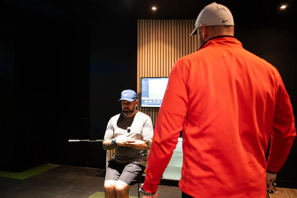 Golfer seated with a club across the chest during a seated rotation test while the coach stands by at Evolution Golf Academy near Derby