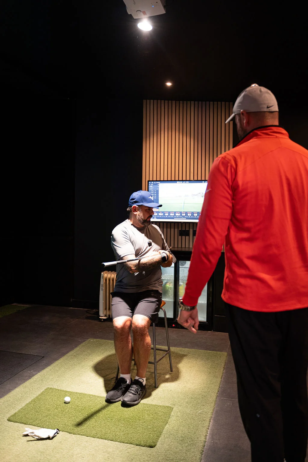 Seated golfer holding a club across the arms on the hitting mat while the coach watches at Evolution Golf Academy near Derby