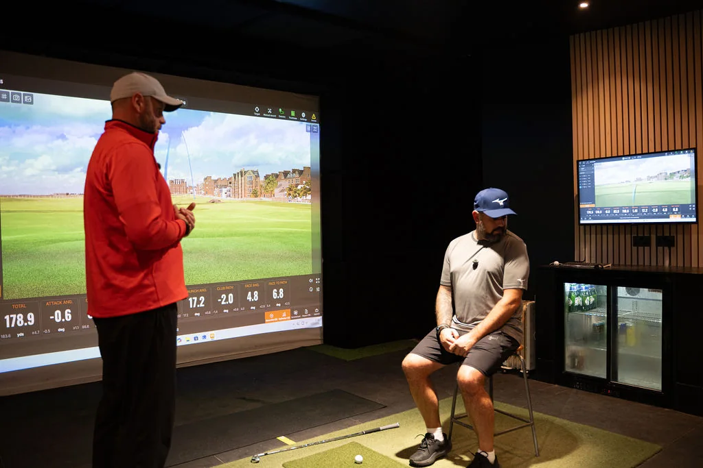 Coach explaining movement results to a seated golfer in front of the simulator screen at Evolution Golf Academy near Derby