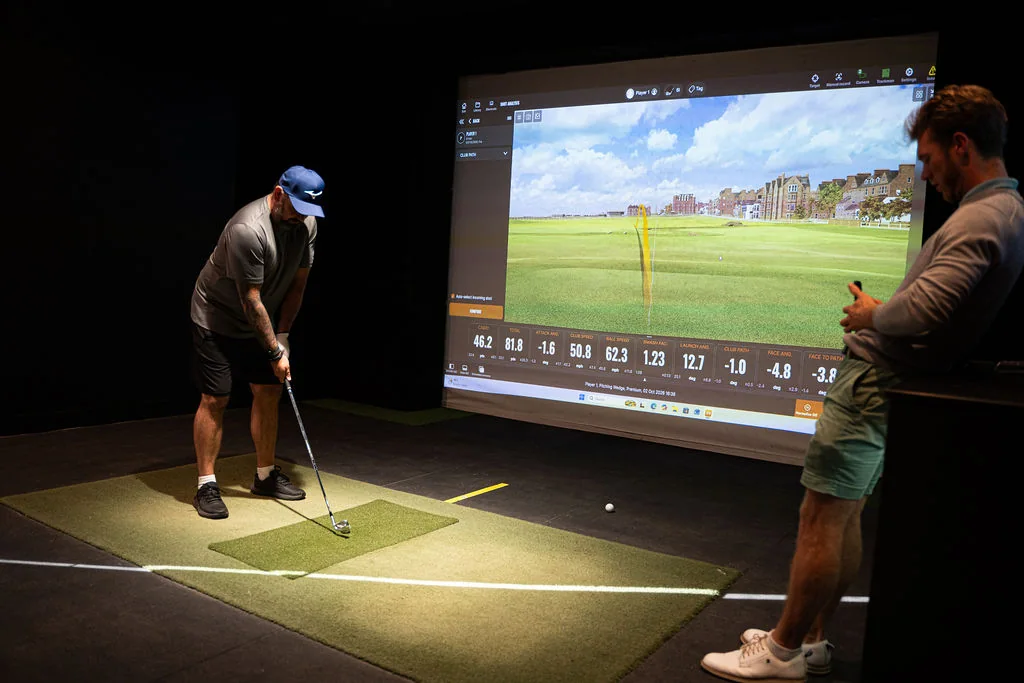 Golfer playing a shot while a coach looks on in the simulator bay at Evolution Golf Academy near Derby