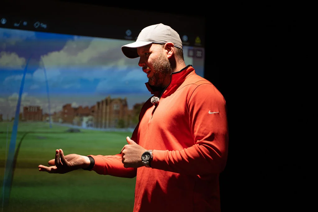 Coach explaining a swing fault with an open hand in front of the simulator screen at Evolution Golf Academy near Derby