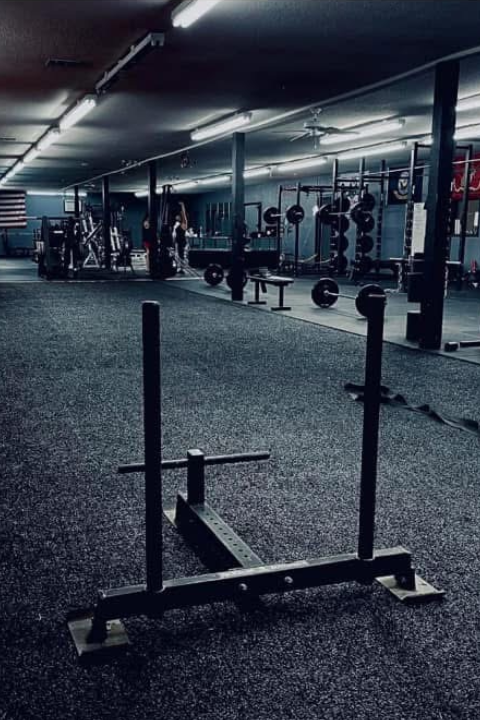 A dimly lit gym with racks of weights, benches, and machines. A red carpet runs along one side, and flags hang on the walls, creating a serious, focused atmosphere.
