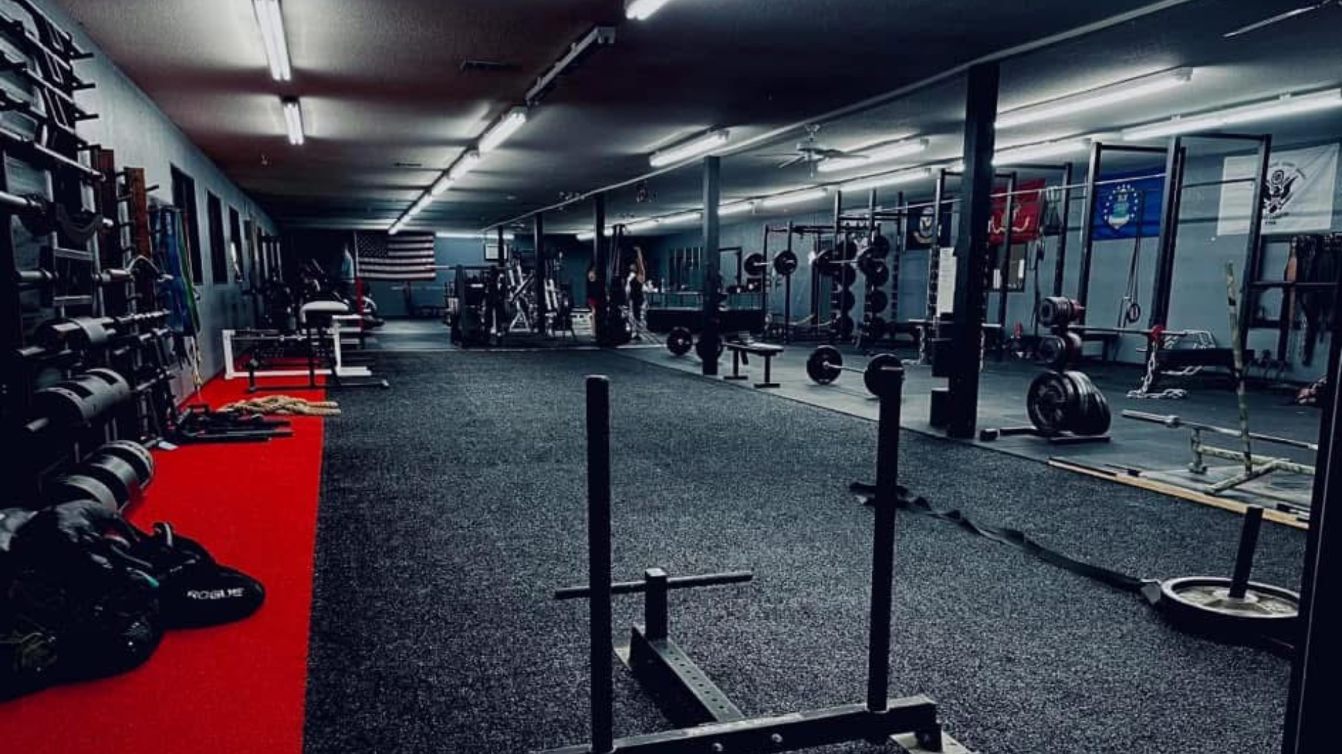 A dimly lit gym with racks of weights, benches, and machines. A red carpet runs along one side, and flags hang on the walls, creating a serious, focused atmosphere.