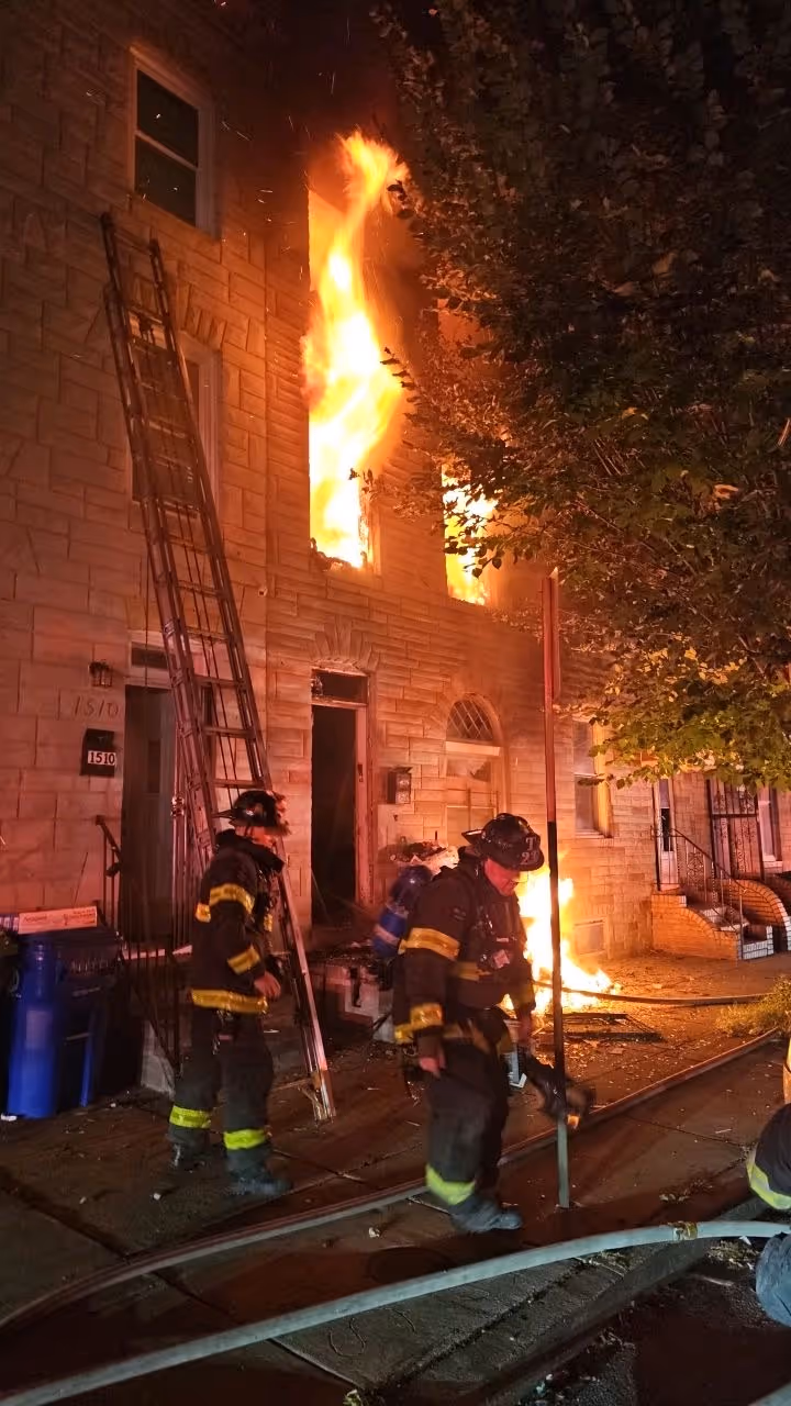 Firefighters stand outside a brick building with flames rising from a lower-floor doorway and upper window, with a ladder leaned against the facade and hoses on the street.