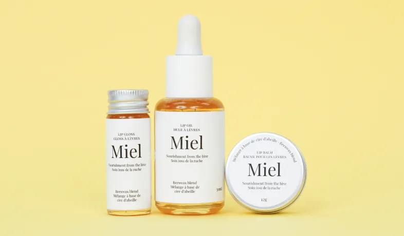 Three Miel lip care products with beeswax blend on a yellow background: gloss, oil, and balm.