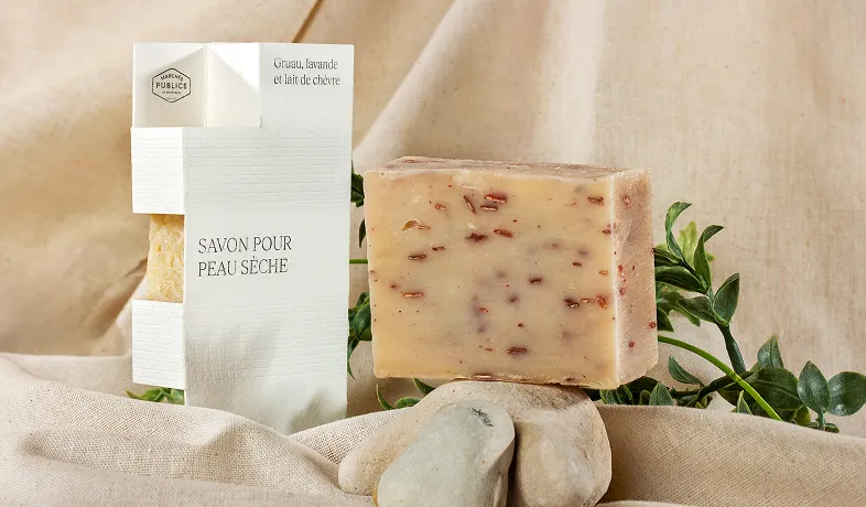 Bar of natural soap with packaging labeled 'Savon Pour Peau Sèche' and herbs on beige fabric background.