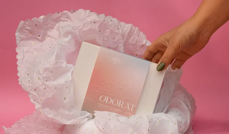 Hand with green nail polish holding a pastel pink and white box labeled ODORAT over tissue paper.