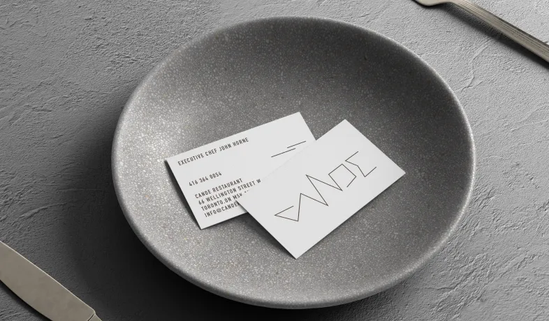 Two restaurant business cards placed in a gray speckled bowl on a textured surface with a knife and spoon nearby.
