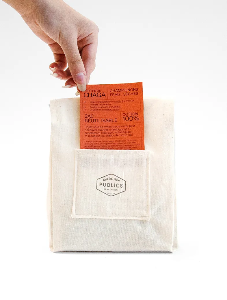 Hand placing an orange packet labeled CHAGA mushrooms into a beige reusable cotton bag with Marchés Publics logo.