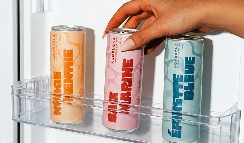 Hand holding pink kombucha can labeled Baie Marine among other cans in a refrigerator door shelf.