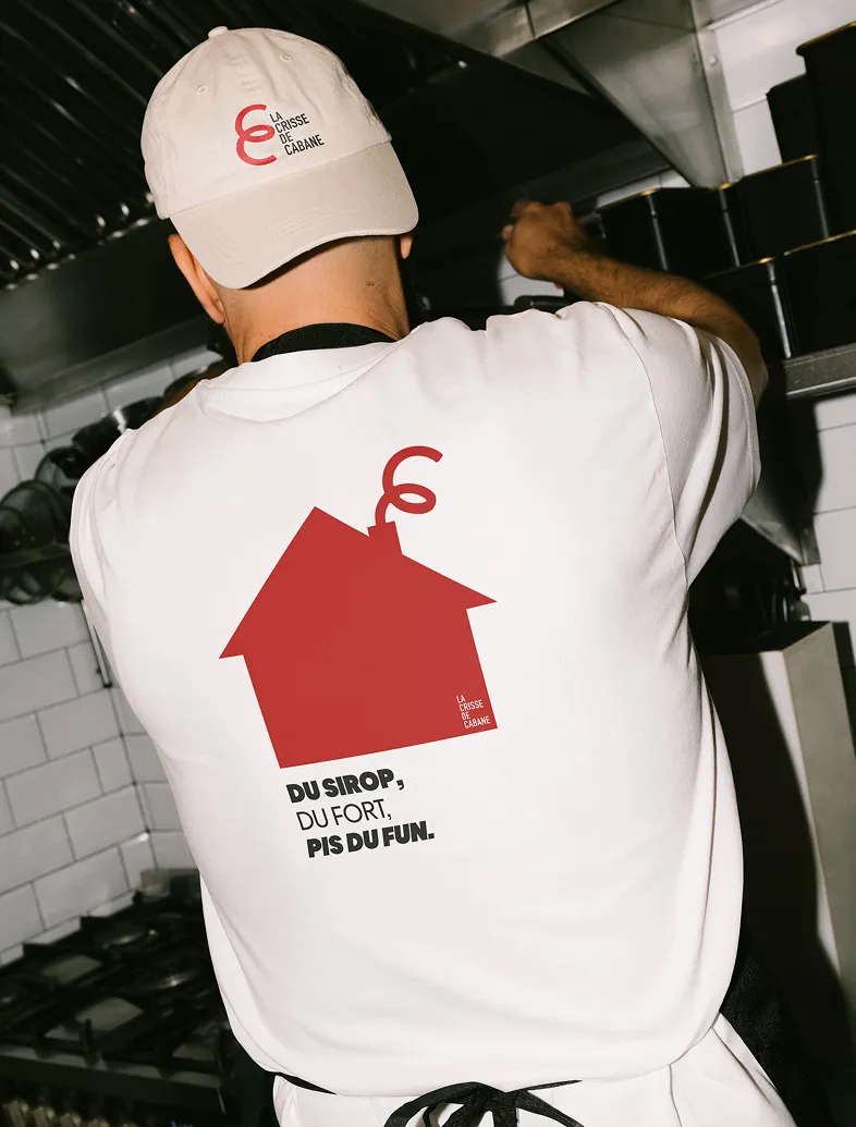 Person wearing white cap and shirt with red house logo and French text in a commercial kitchen.
