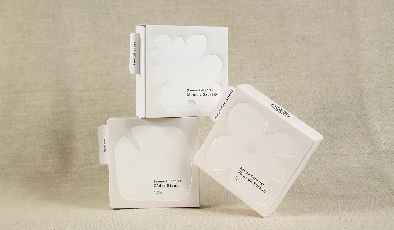 Three white minimalist boxes of body balm with floral and leaf cutout designs on beige backdrop.