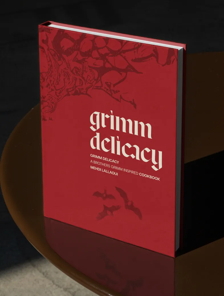Red cookbook titled Grimm Delicacy with bats and tree design on brown reflective surface.
