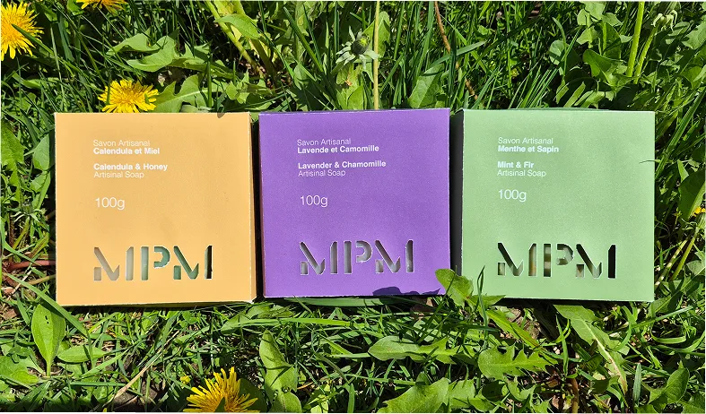 Three MPM artisanal soap boxes—calendula and honey, lavender and chamomile, mint and fir—on grass with dandelions.