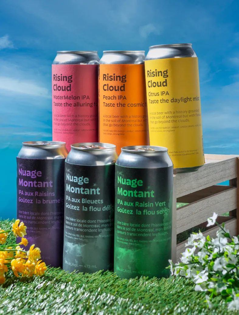 Six colorful Rising Cloud and Nuage Montant IPA cans on grass and wooden crate under blue sky.