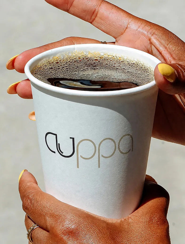 Hands with yellow nail polish holding a white cup of black coffee labeled 'cuppa'.