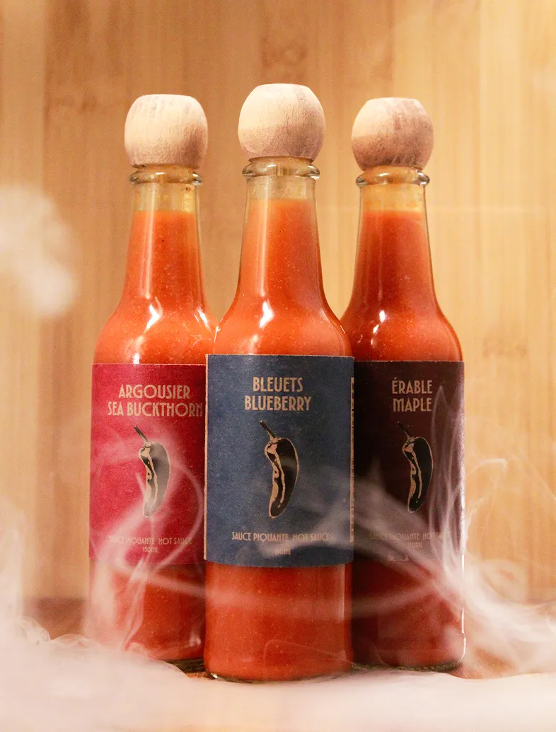 Three glass bottles of hot sauce with wooden stoppers labeled Sea Buckthorn, Blueberry, and Maple.