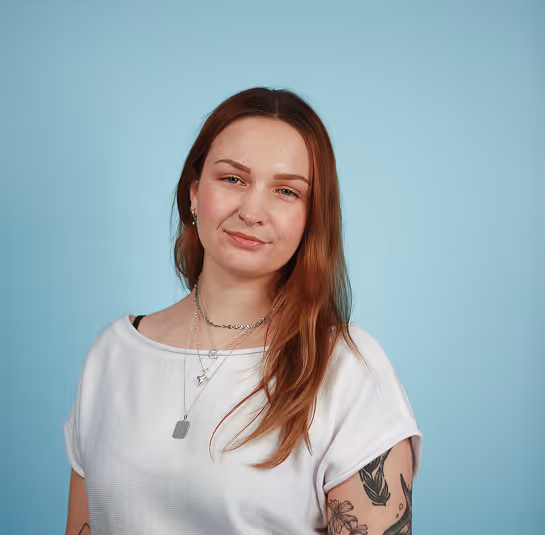 Woman with long red hair wearing white top and layered necklaces, with tattoos on her arm, against blue background.