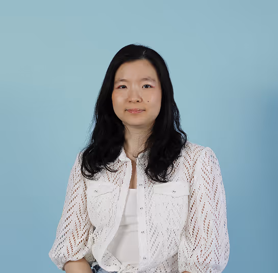 Woman with long black hair wearing a white patterned blouse and white top against a light blue background.