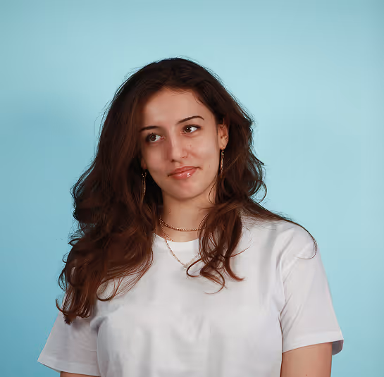 Woman with long brown hair wearing a white t-shirt and gold jewelry against a light blue background.