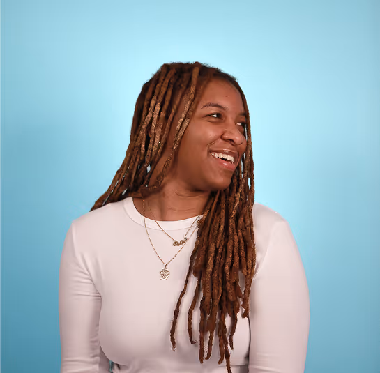 Woman with long brown dreadlocks wearing white top and gold necklaces smiling against a blue background.
