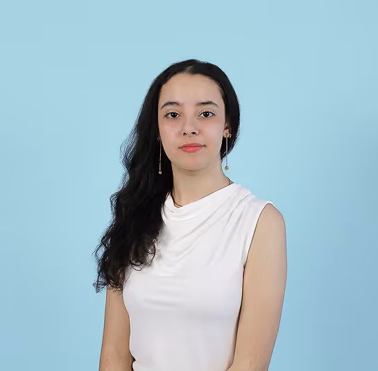 Young woman with long dark hair wearing white sleeveless top, posing against light blue background.