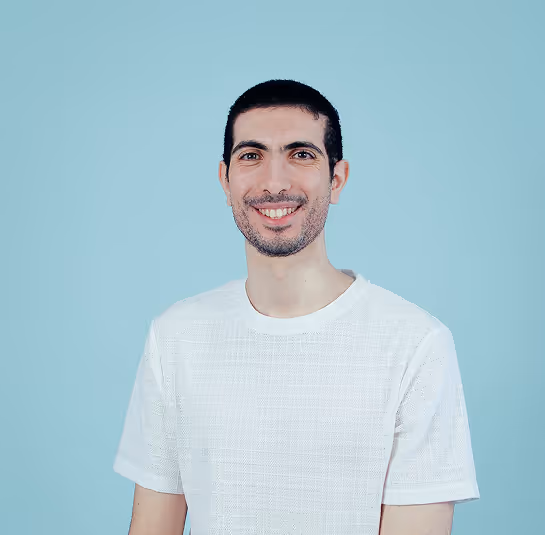 Smiling man with short dark hair and beard wearing a white shirt against a light blue background.