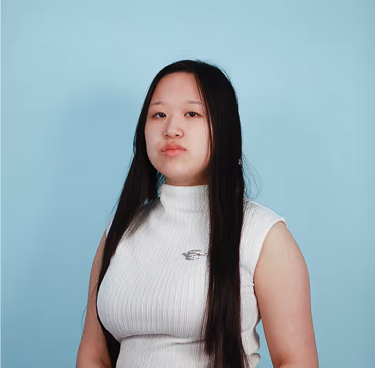 Woman with long black hair wearing a white sleeveless top standing against a light blue background.