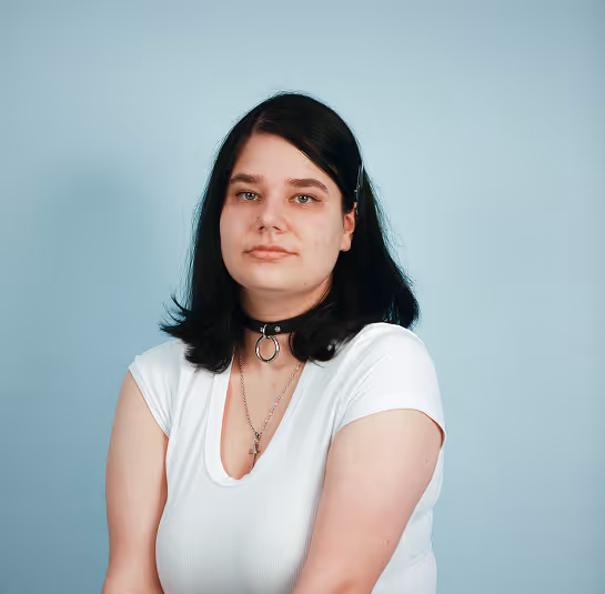 Young woman with medium-length dark hair wearing a white shirt and black choker against a plain blue background.