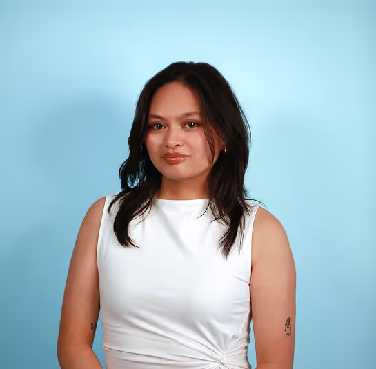 Woman with dark hair wearing a white sleeveless top against a light blue background.