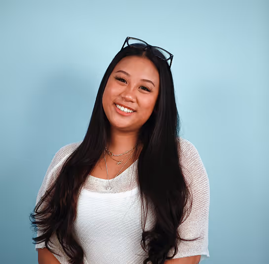 Smiling woman with long black hair wearing a white top and glasses resting on her head against a blue background.