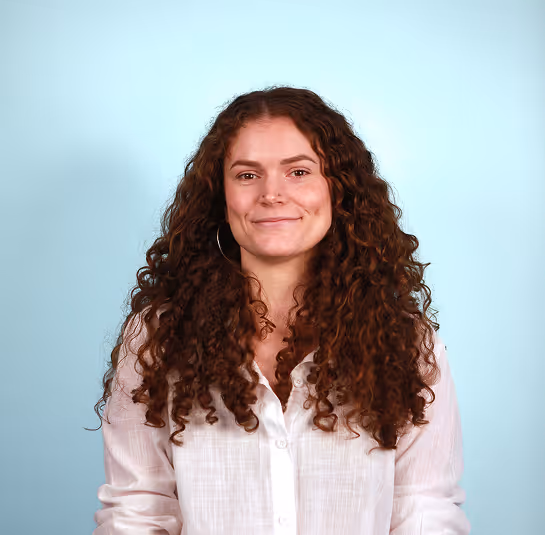 Smiling woman with long curly hair wearing a white button-up shirt against a light blue background.