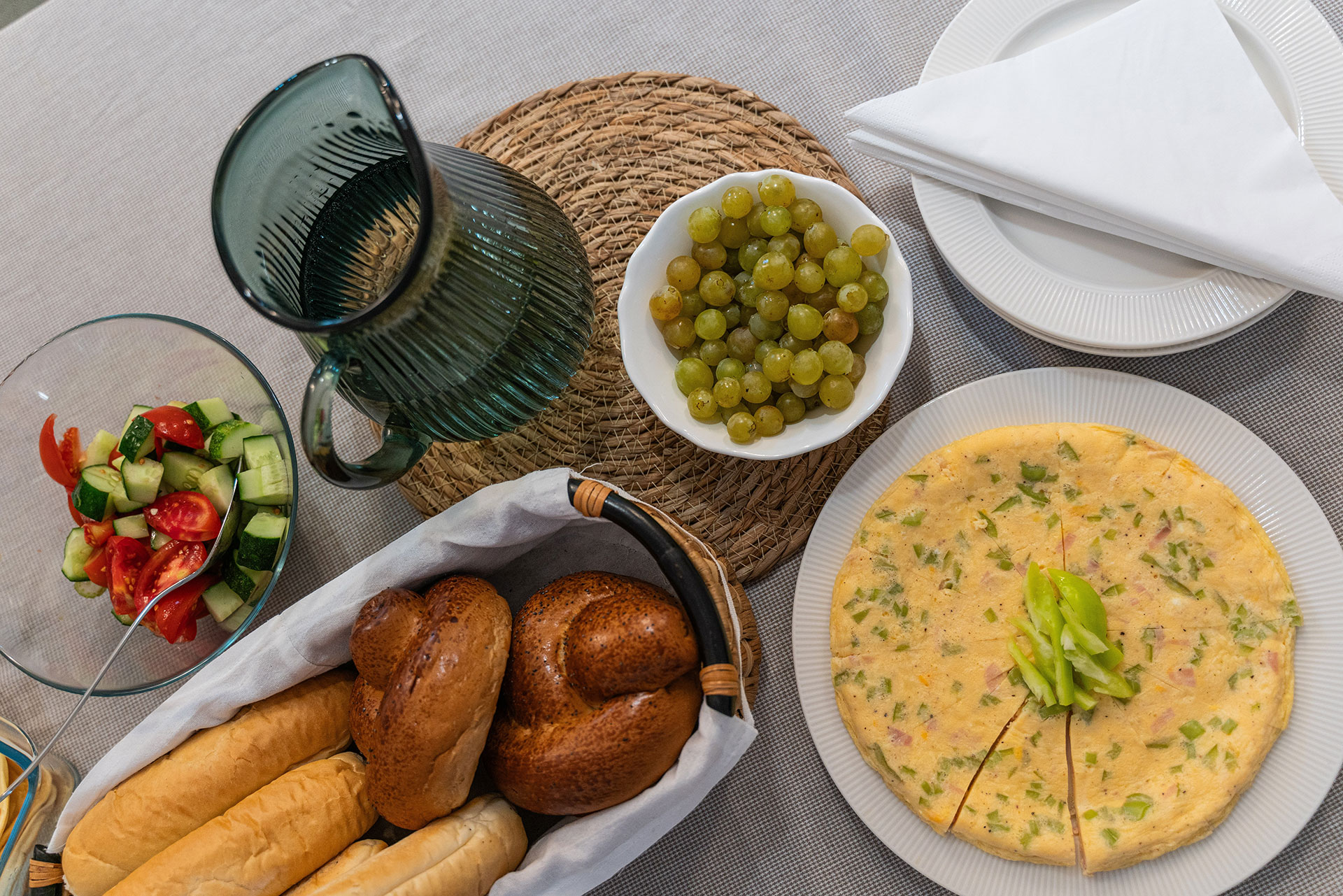 Table with basket of rolls, green grapes, omelet garnished with green peppers, salad, green pitcher, and plates with napkins.
