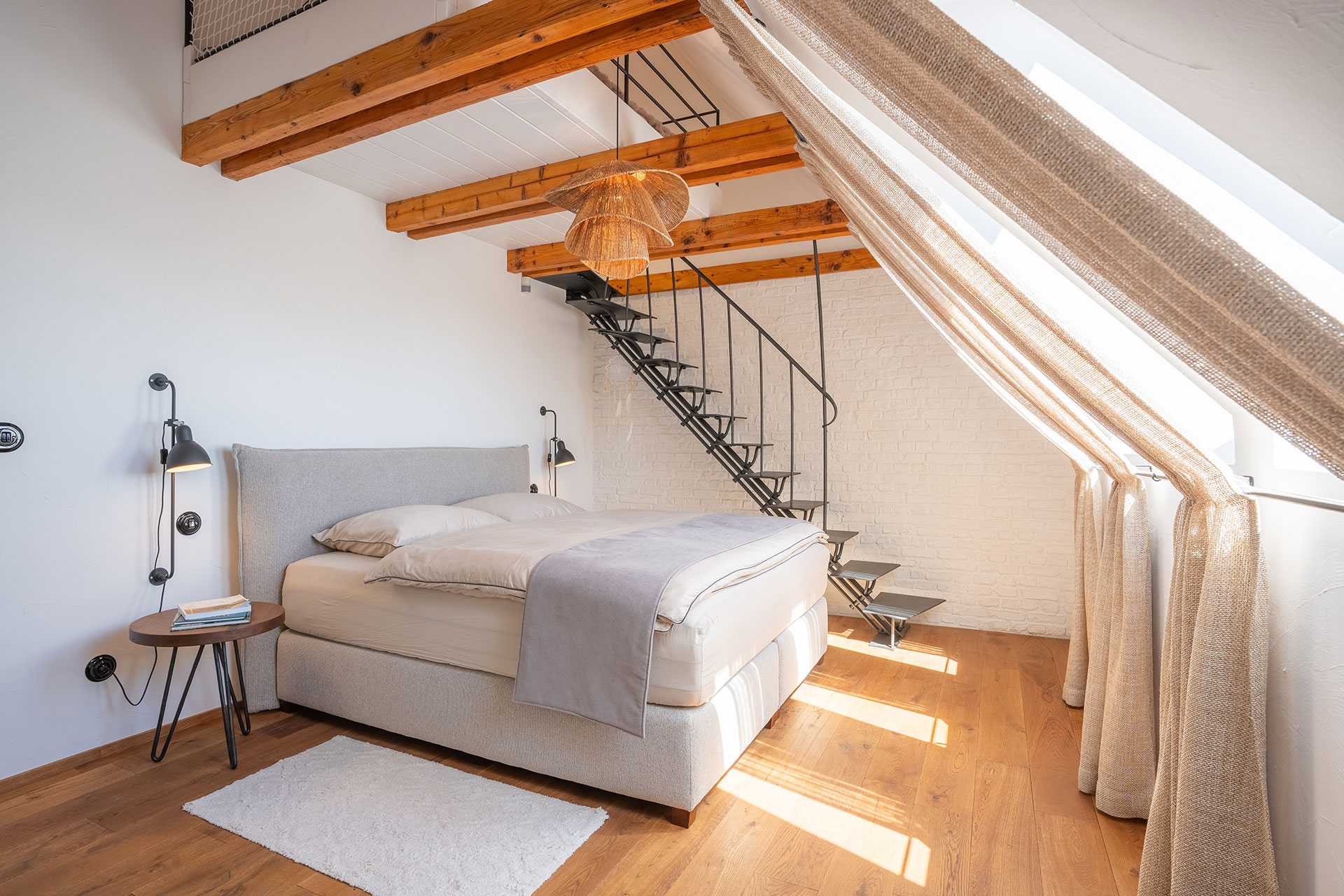 Sunlit modern bedroom with wooden beams, light bed, side table, and black metal staircase.
