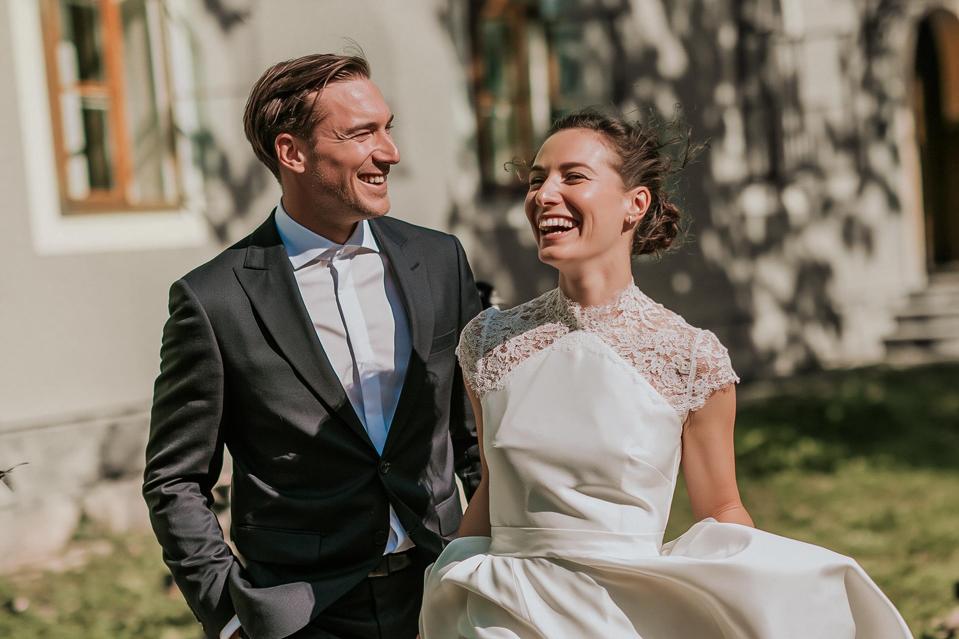 Smiling bride in lace wedding dress and groom in black suit standing outside in sunlight.