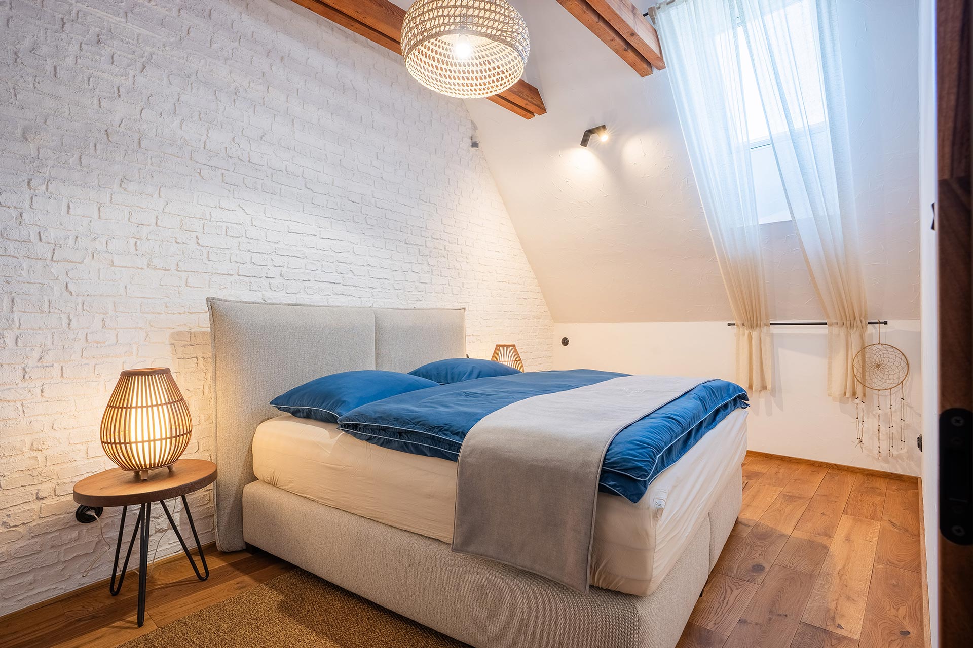 Cozy bedroom with beige upholstered bed, blue bedding, wooden floor, wicker lamps, and white brick wall.
