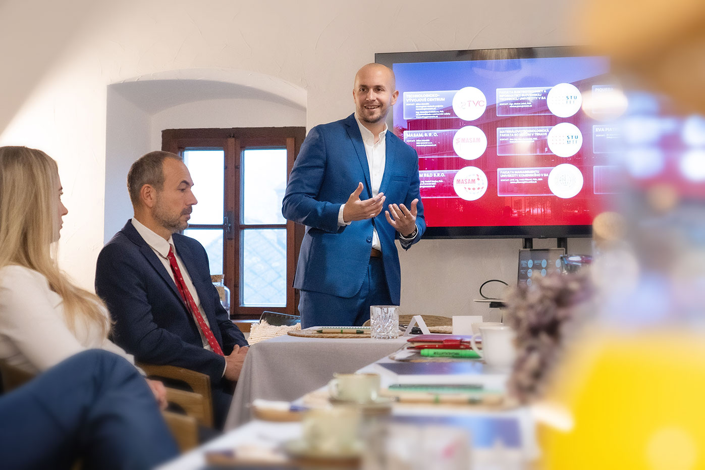 Man in blue suit giving presentation to seated colleagues in a meeting room.