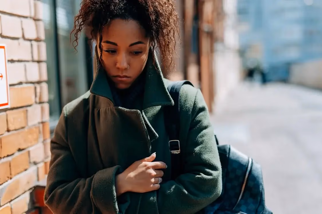 A woman with curly hair stands in an alleyway beside a brick wall with a red sign, wearing a green coat and holding a dark backpack.