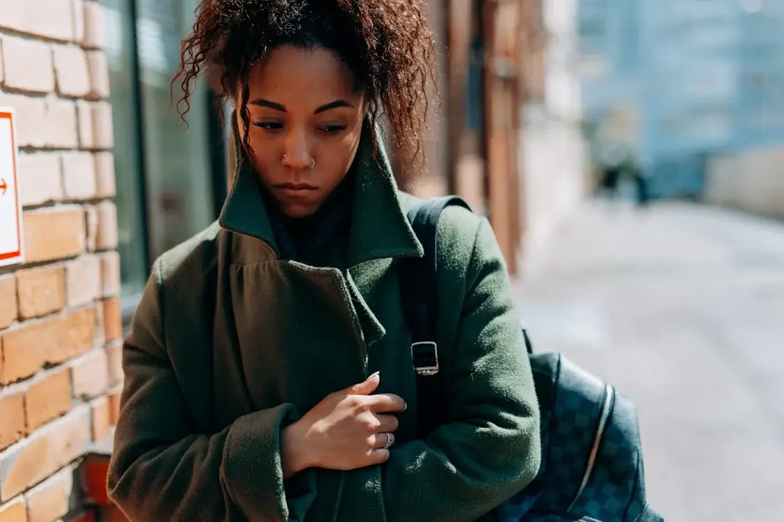 A woman with curly hair stands in an alleyway beside a brick wall with a red sign, wearing a green coat and holding a dark backpack.