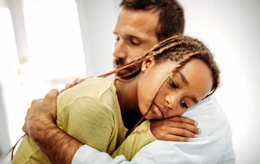 A man embraces a young girl with braided hair as she rests her head on his shoulder.