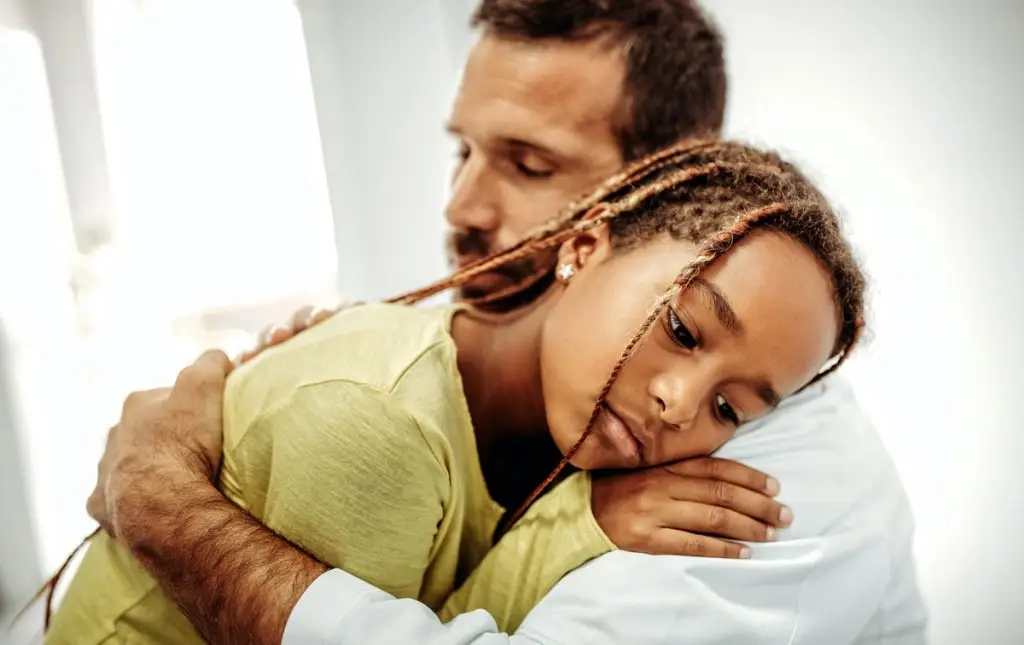 A man embraces a young girl with braided hair as she rests her head on his shoulder.