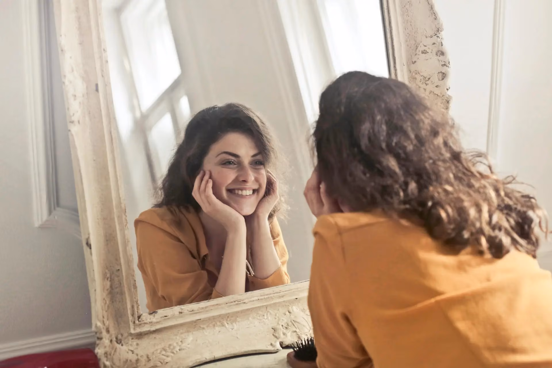 A smiling woman holds her cheeks while looking at her reflection in a vintage mirror near a sunlit window.
