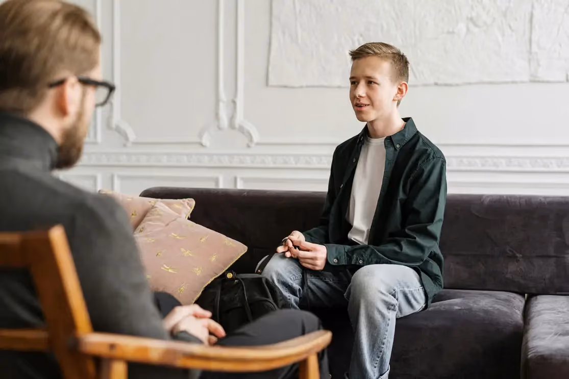 A young man sits on a dark couch speaking to an adult in a wooden chair during a conversation.
