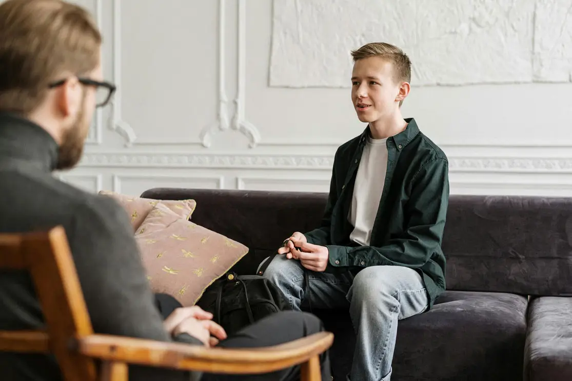 A young man sits on a dark couch speaking to an adult in a wooden chair during a conversation.