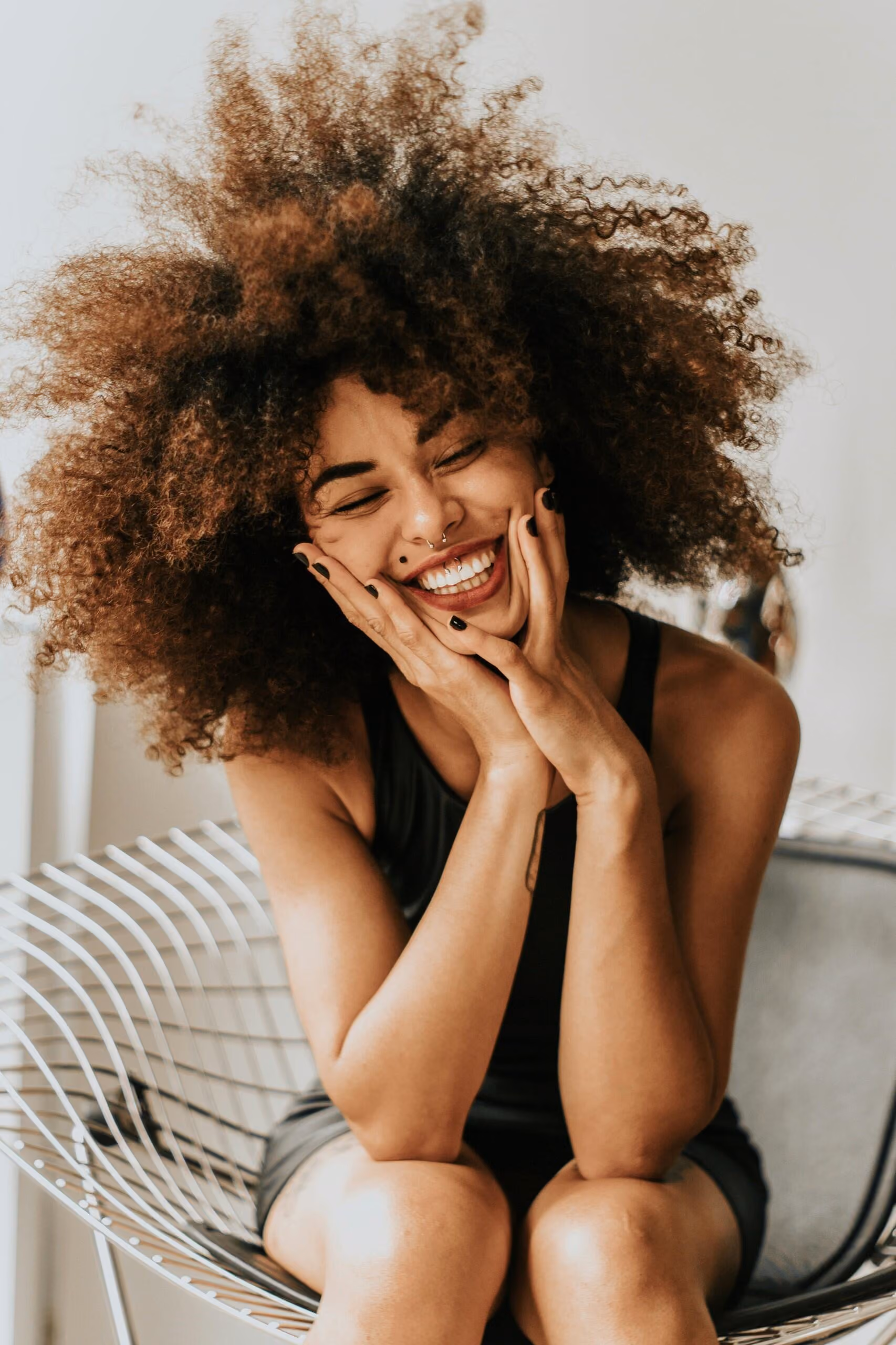 A smiling woman with voluminous curly hair holds her cheeks with both hands while seated on a modern wire chair.