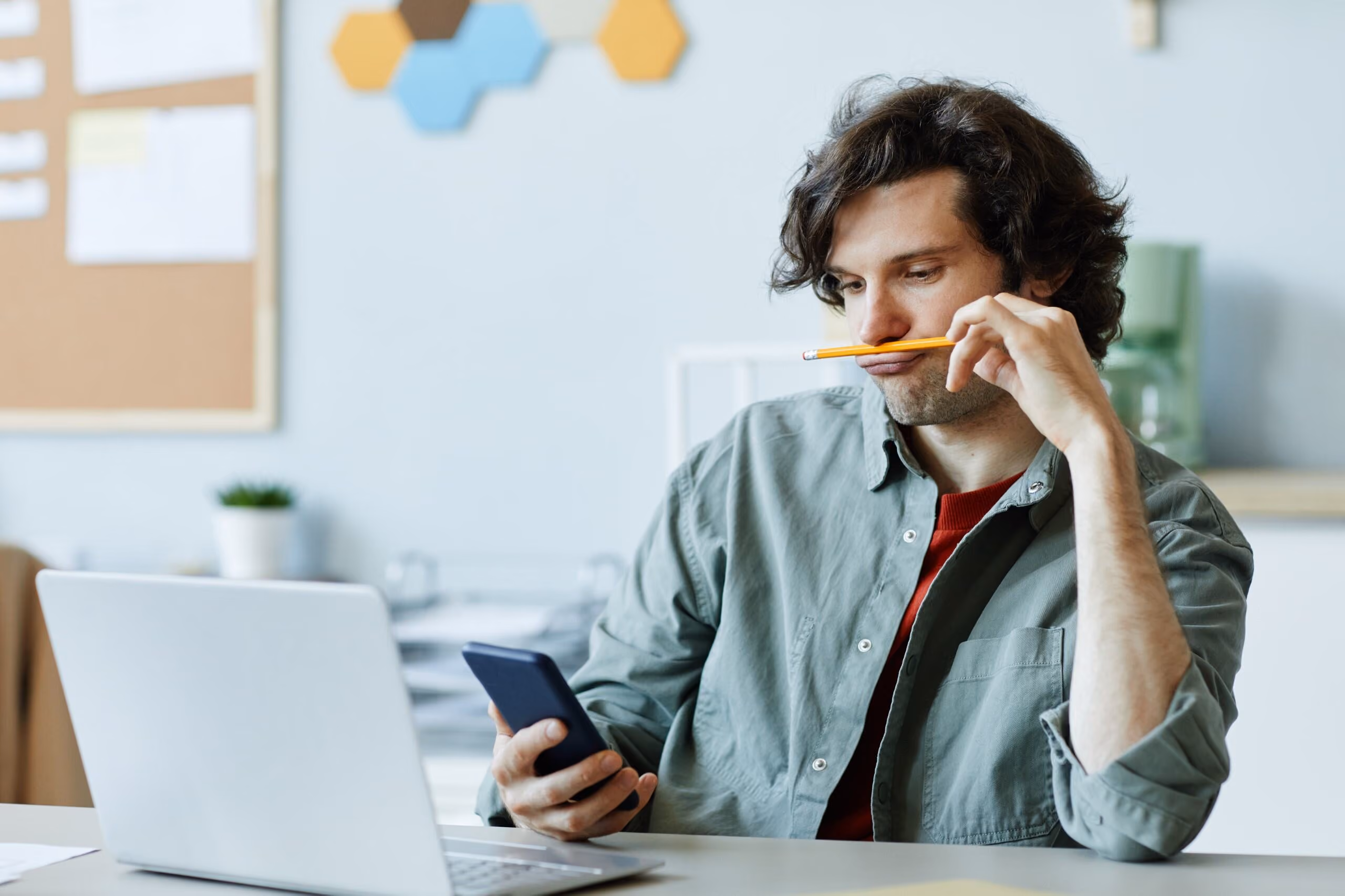 Man holds a smartphone while looking at a laptop with a pencil near his mouth.