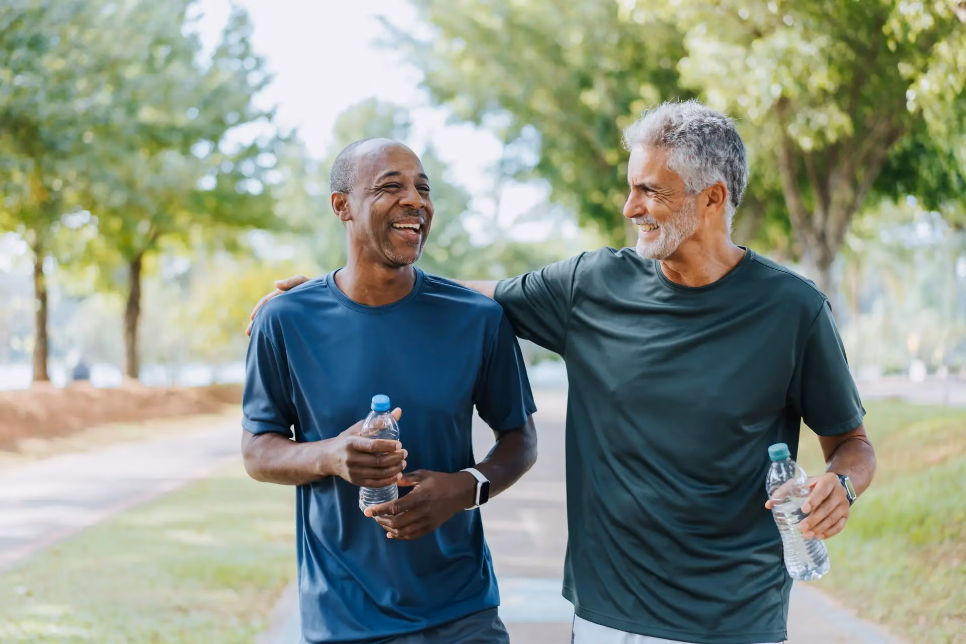 Two men walk together outdoors, each holding a water bottle and smiling.